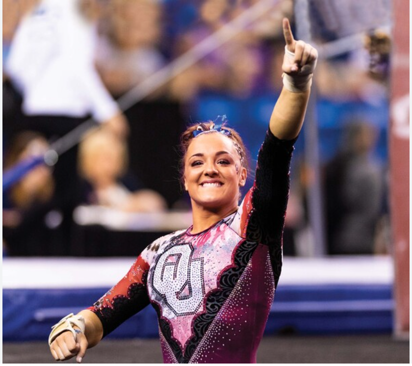 Maggie Nichols smiles and holds her left arm aloft, pointing one finger at something off camera in celebration.